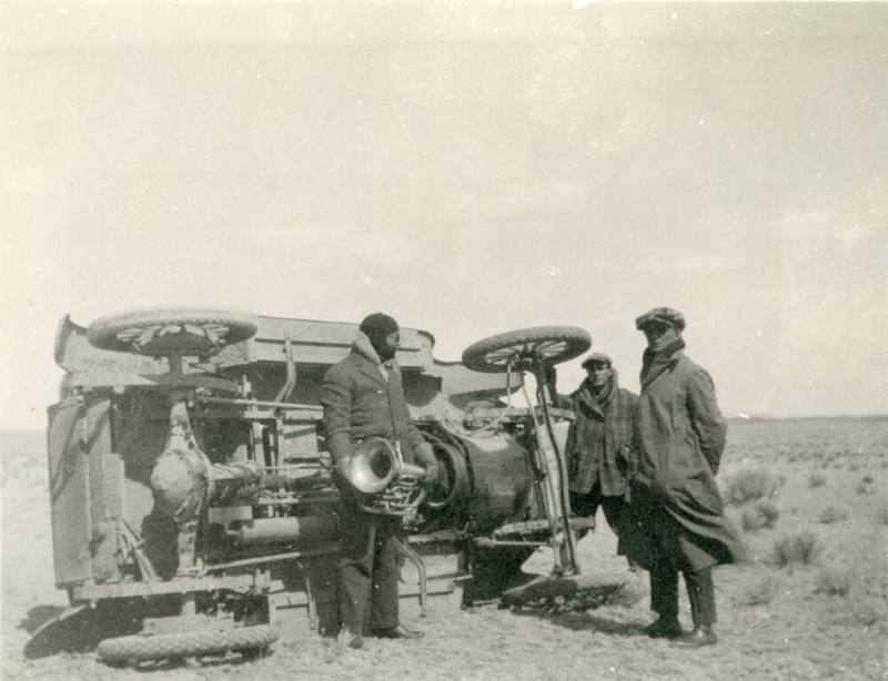 Image of 3 men standing in front of an overturned car in the desert
